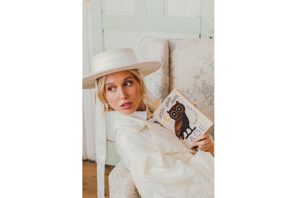 Woman in white hat sat on a chair holding white clutch with owl design