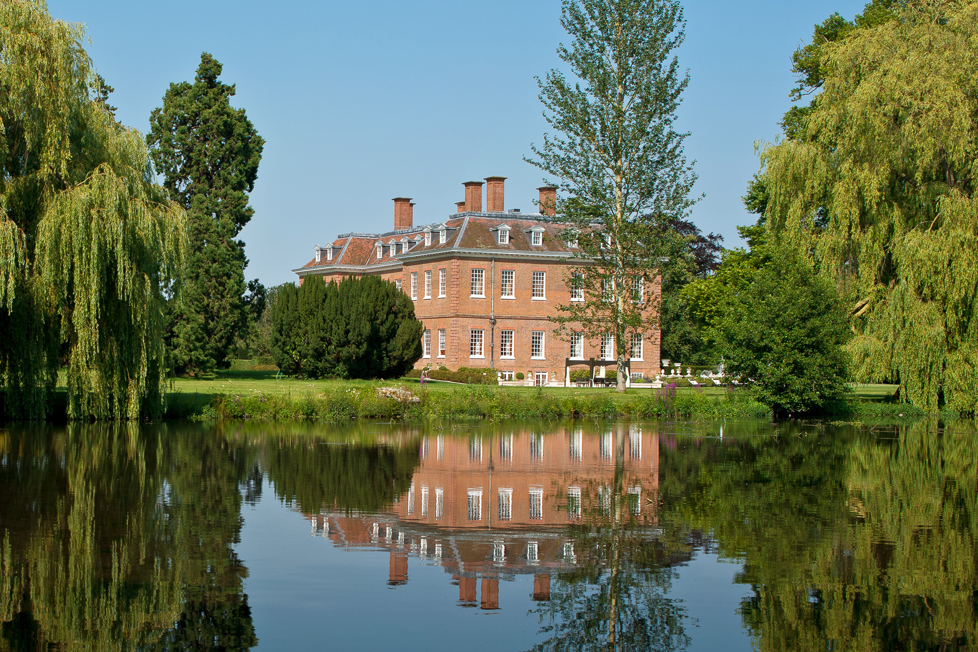 Red brick mansion with a trees surrounding and a lake in front. 