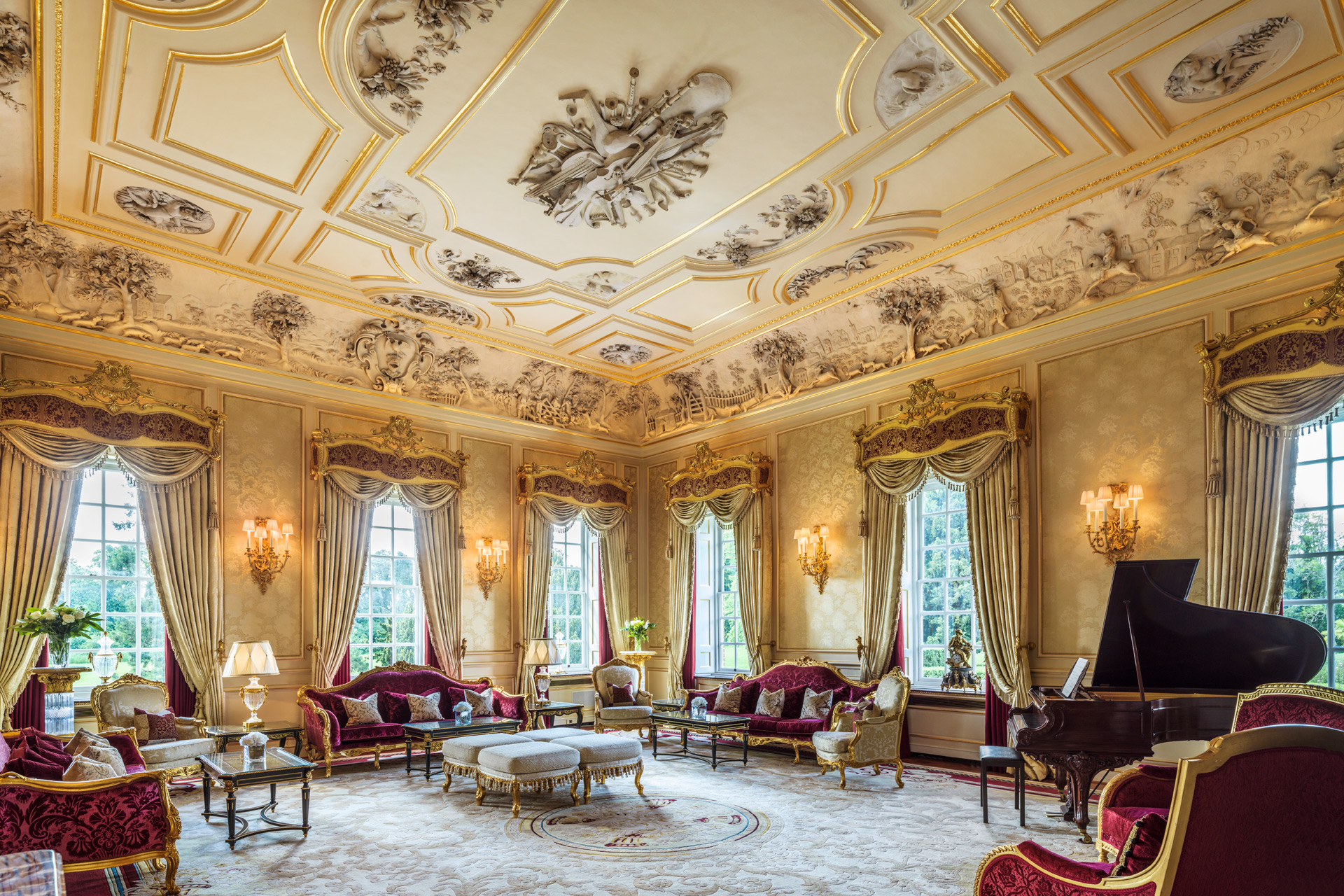 Music room with red plush seating, Persian rug, grand piano and ornate ceiling plaster.