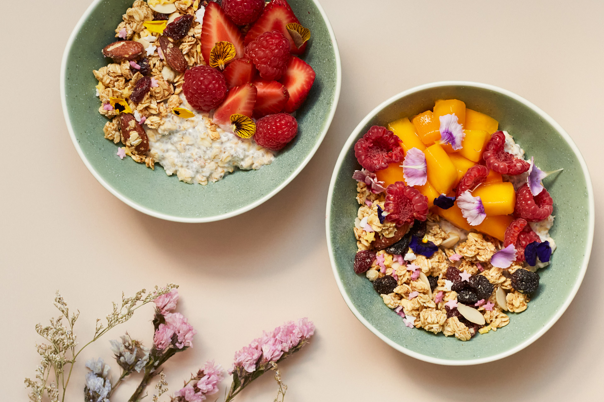 Granola bowls with summer fruits, on a light pink surface with sprigs of dried flowers.