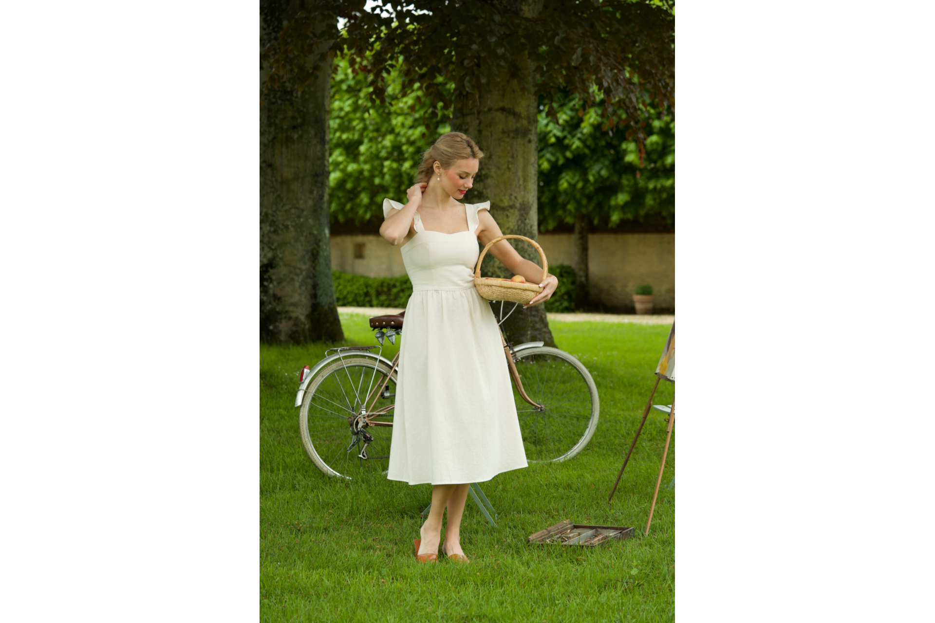 Woman stood on grass next to bike, wearing white dress
