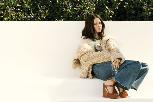 Hayley Atwell sits in a fluffy coat, blue jeans and brown strappy wedges