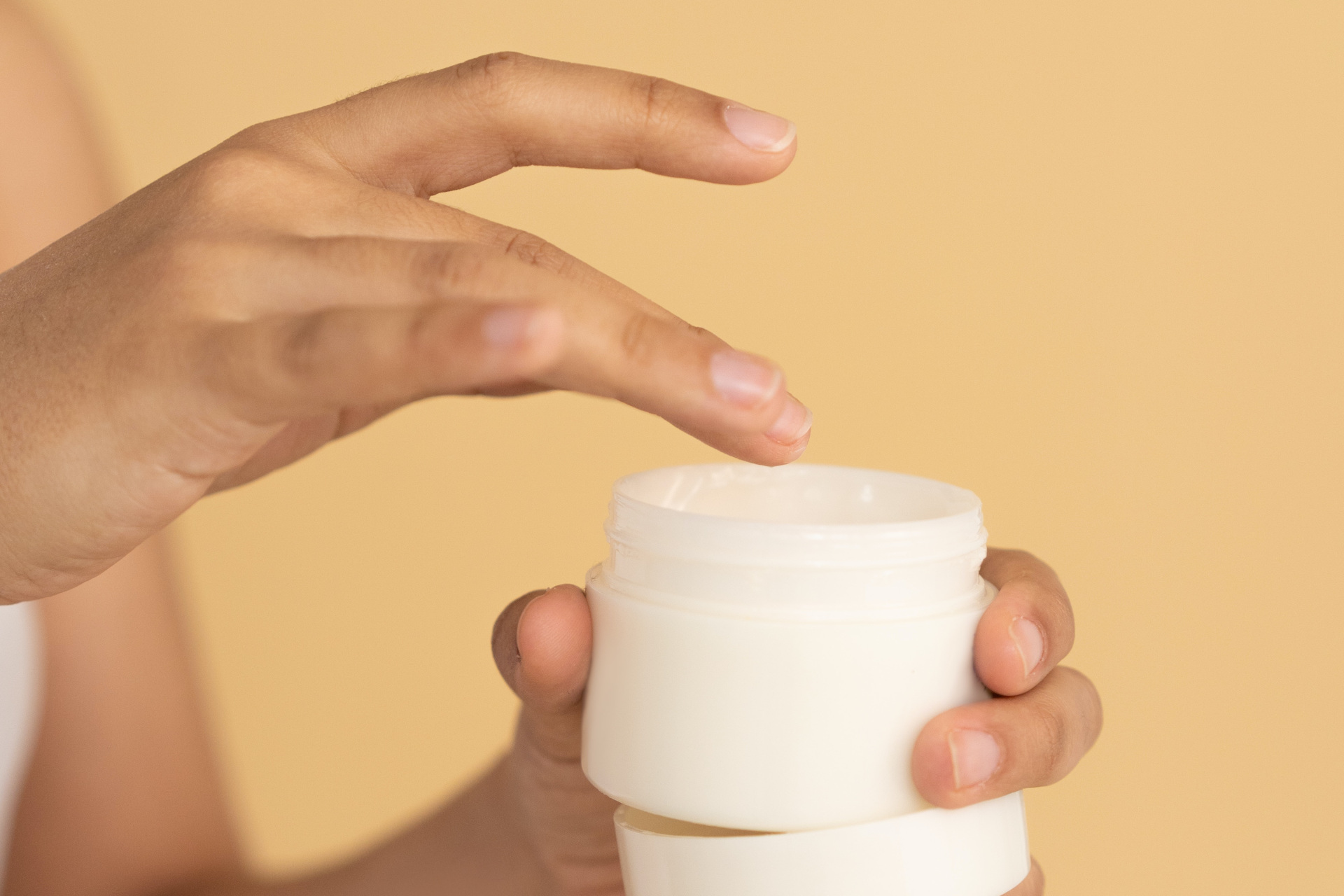 Hand gathering skin cream from white pot on yellow background